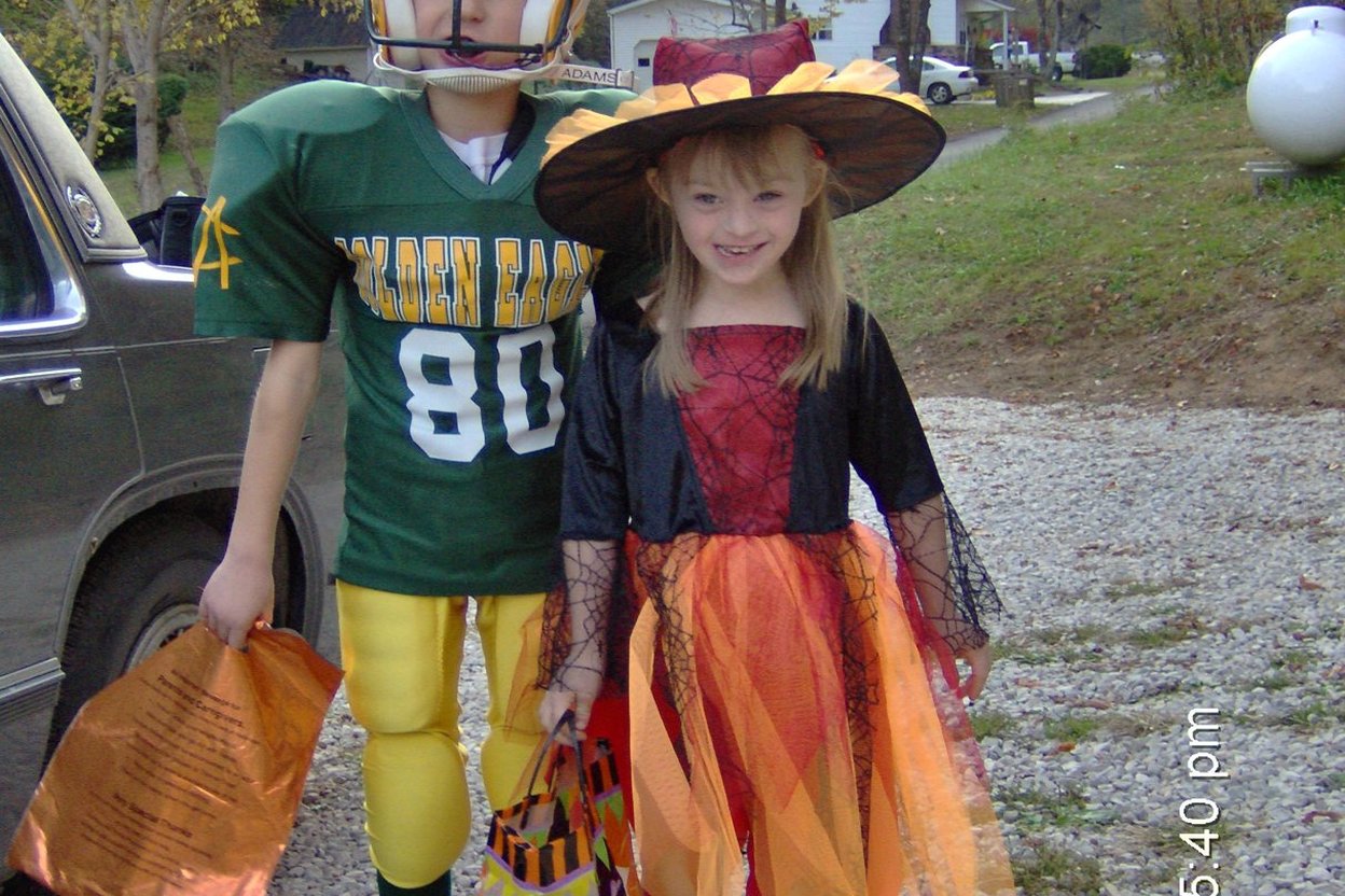 Two children in Halloween costumes standing on a front porch at dusk, holding plastic pumpkin treat buckets.