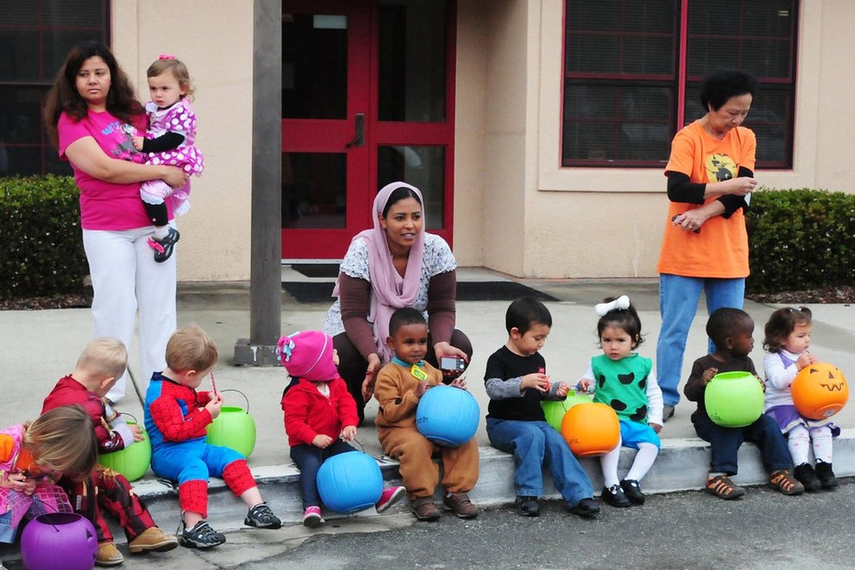 Two parents walking beside a young child in a costume on a residential street at dusk, carrying a trick-or-treat bag.