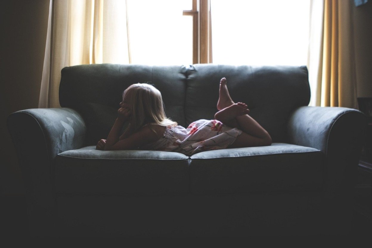 A parent sitting quietly beside a young child on a sofa in a warm living room, with a backpack resting by the front door.
