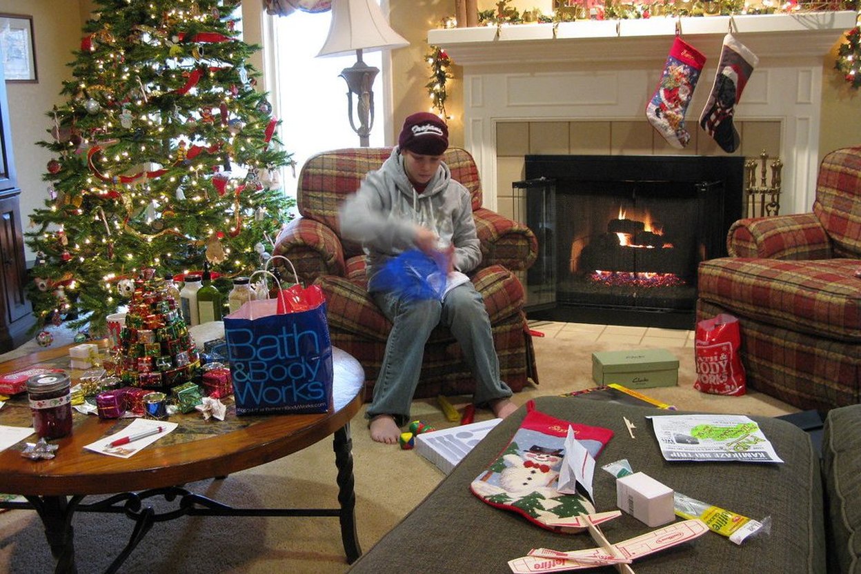 Teenager unwrapping Christmas gifts by fireplace with decorated tree and stockings.