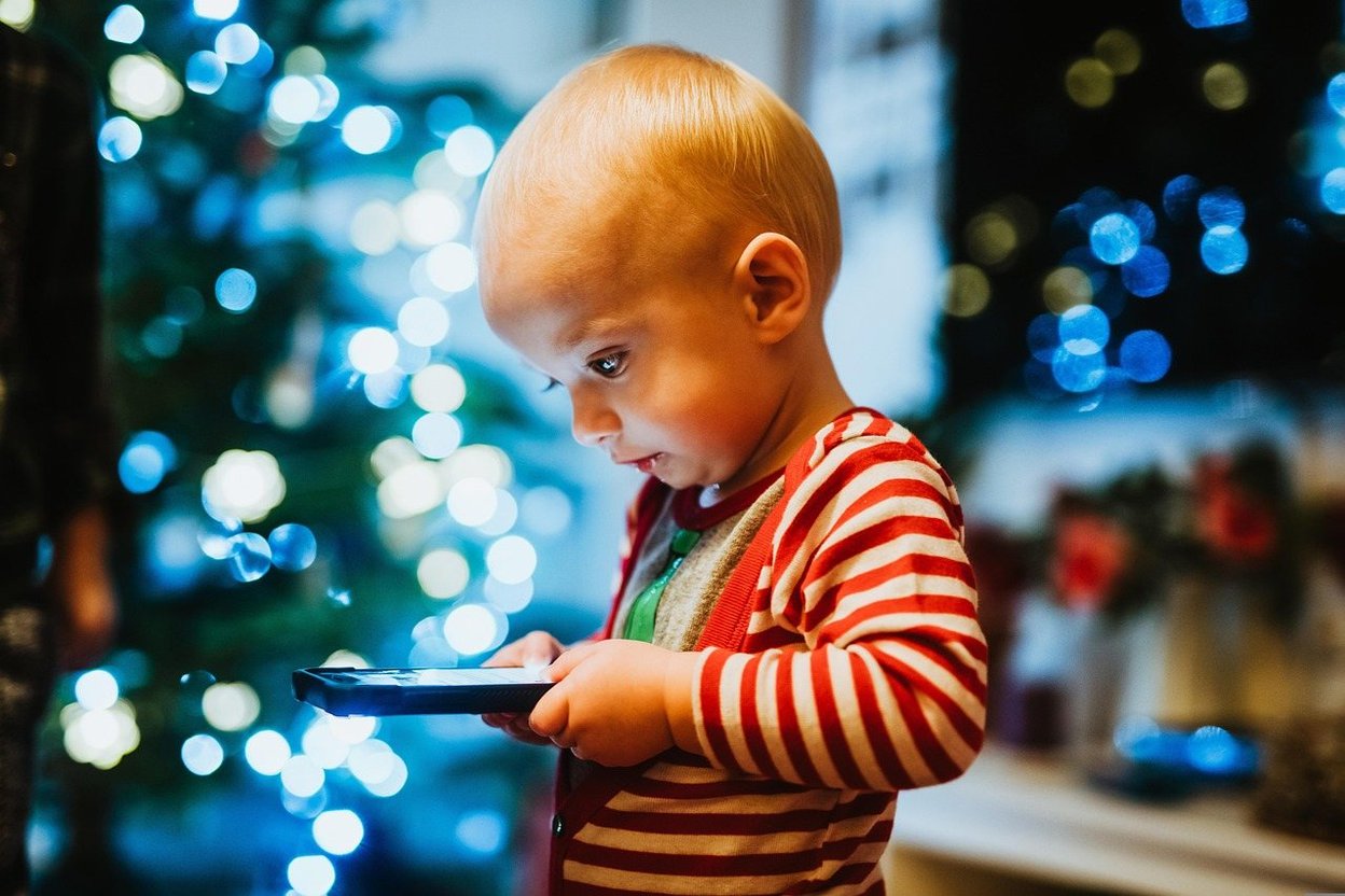 Toddler in red stripes holding tablet, focused against blurred Christmas lights.