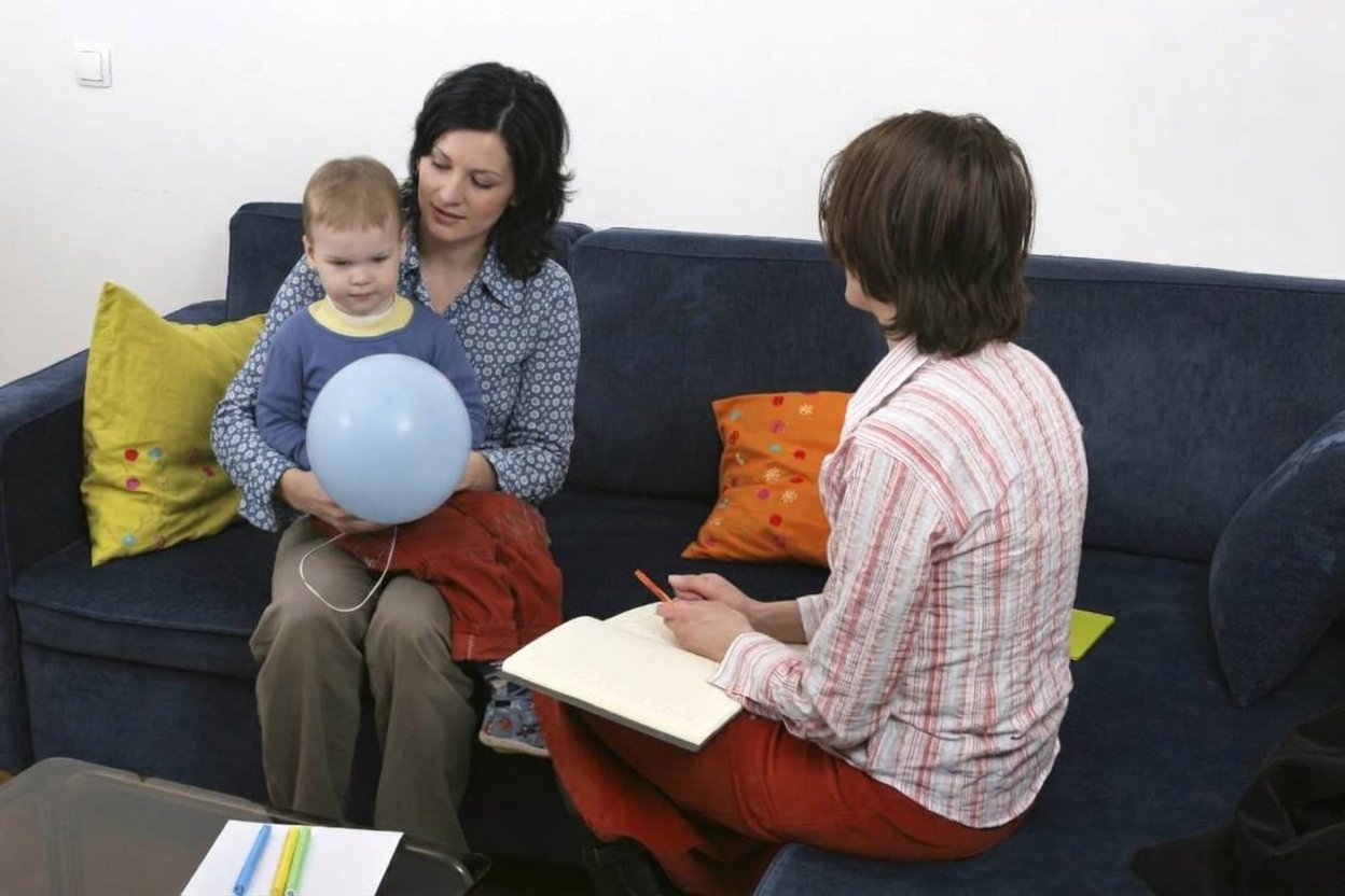 A parent and young child reading together at a table in a family services center, with a supervisor seated quietly nearby.