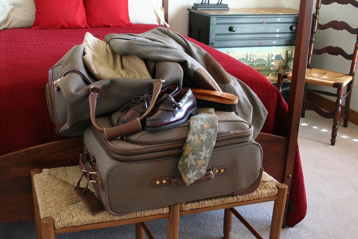 A parent and young child packing a small suitcase together in a bedroom, with a closed moving box nearby.