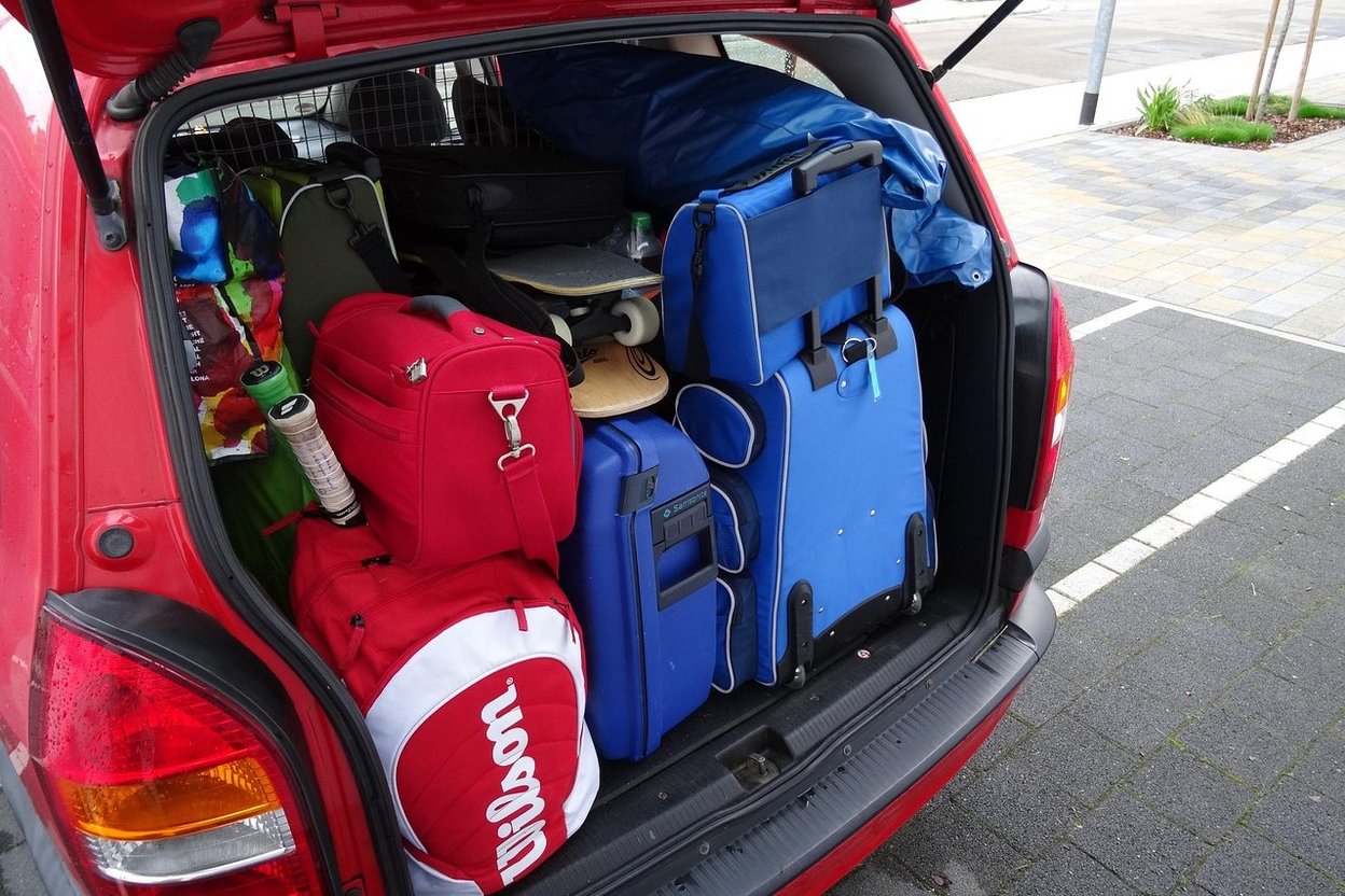 A parent loading suitcases into the trunk of a car while a young child holds a stuffed animal and waits beside them.