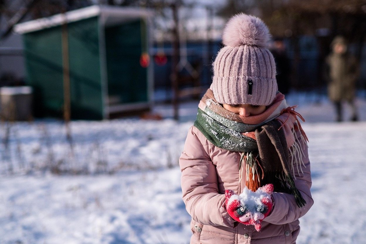 Child in winter gear holding snow, wearing pink hat and scarf outdoors.