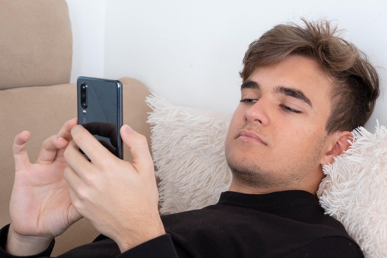 A teenager sitting on a living room couch looking at a smartphone while a parent sits in a chair across the room, both in a quiet home