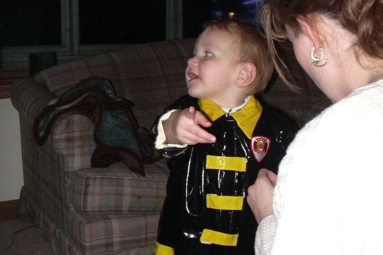 A child in a homemade cardboard costume holds a candy bucket at a front door while a parent stands beside them.
